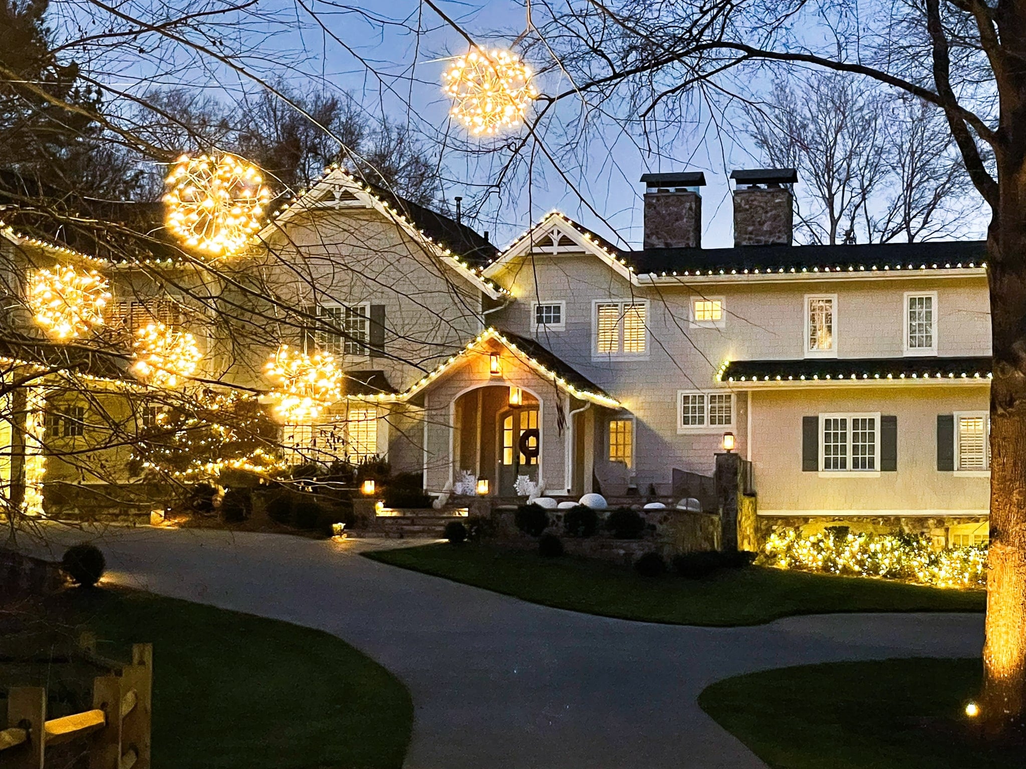 A large house with warm white holiday lights outlining the rooflines, with starburst-shaped ornaments hanging from a nearby tree.
